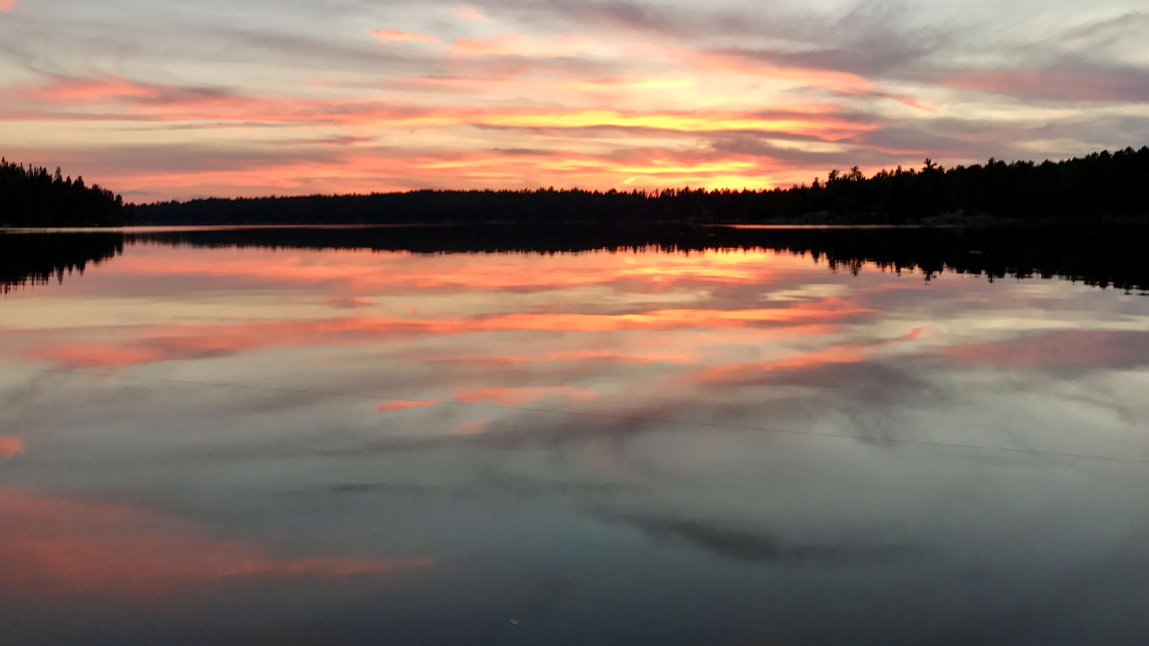 A vibrant orange and pink sunset reflected in the perfectly calm water of a remote Canadian lake lined with pine trees.