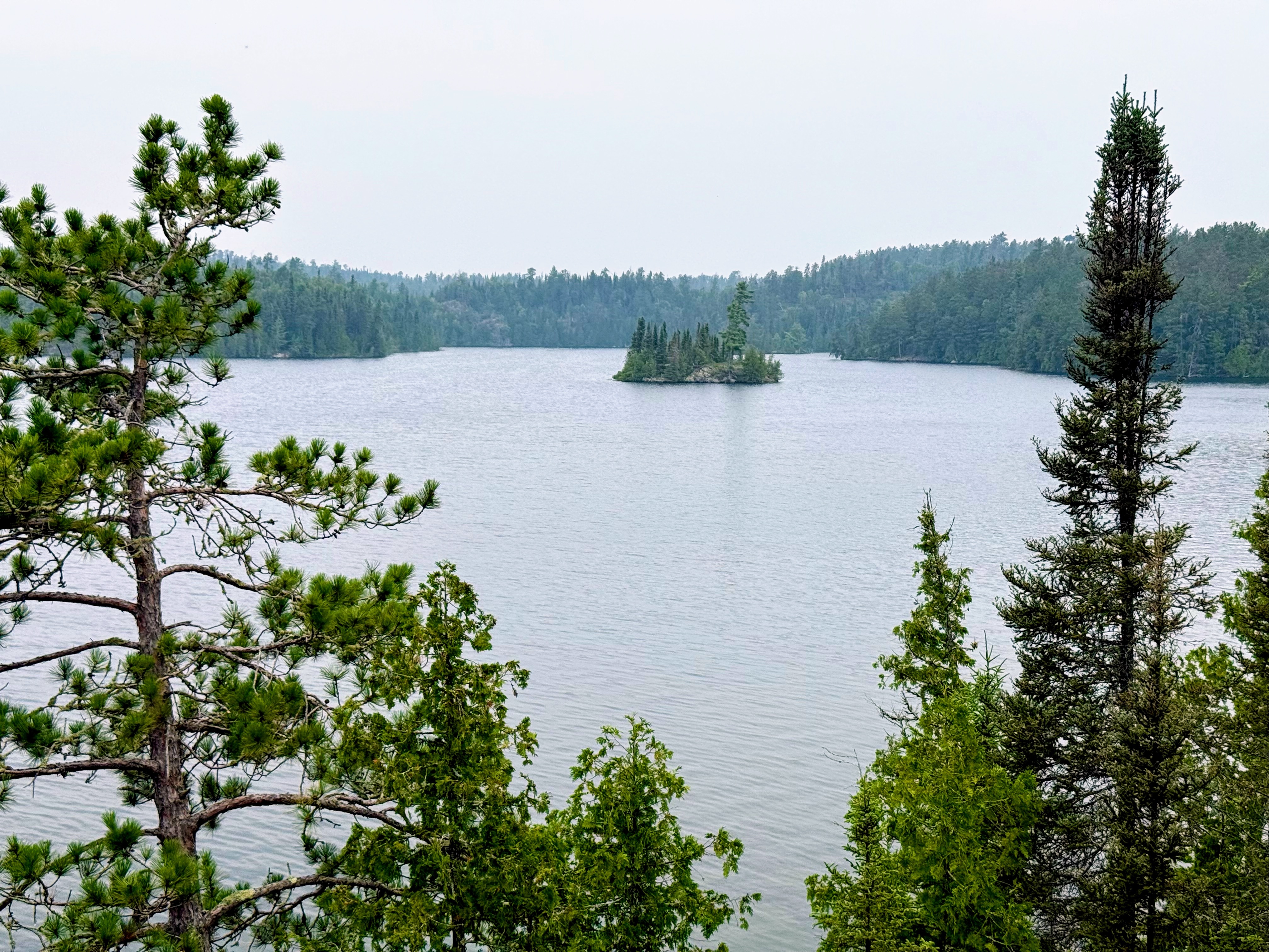 A wide lake surrounded by evergreen trees with a small forested island in the center under a cloudy sky.