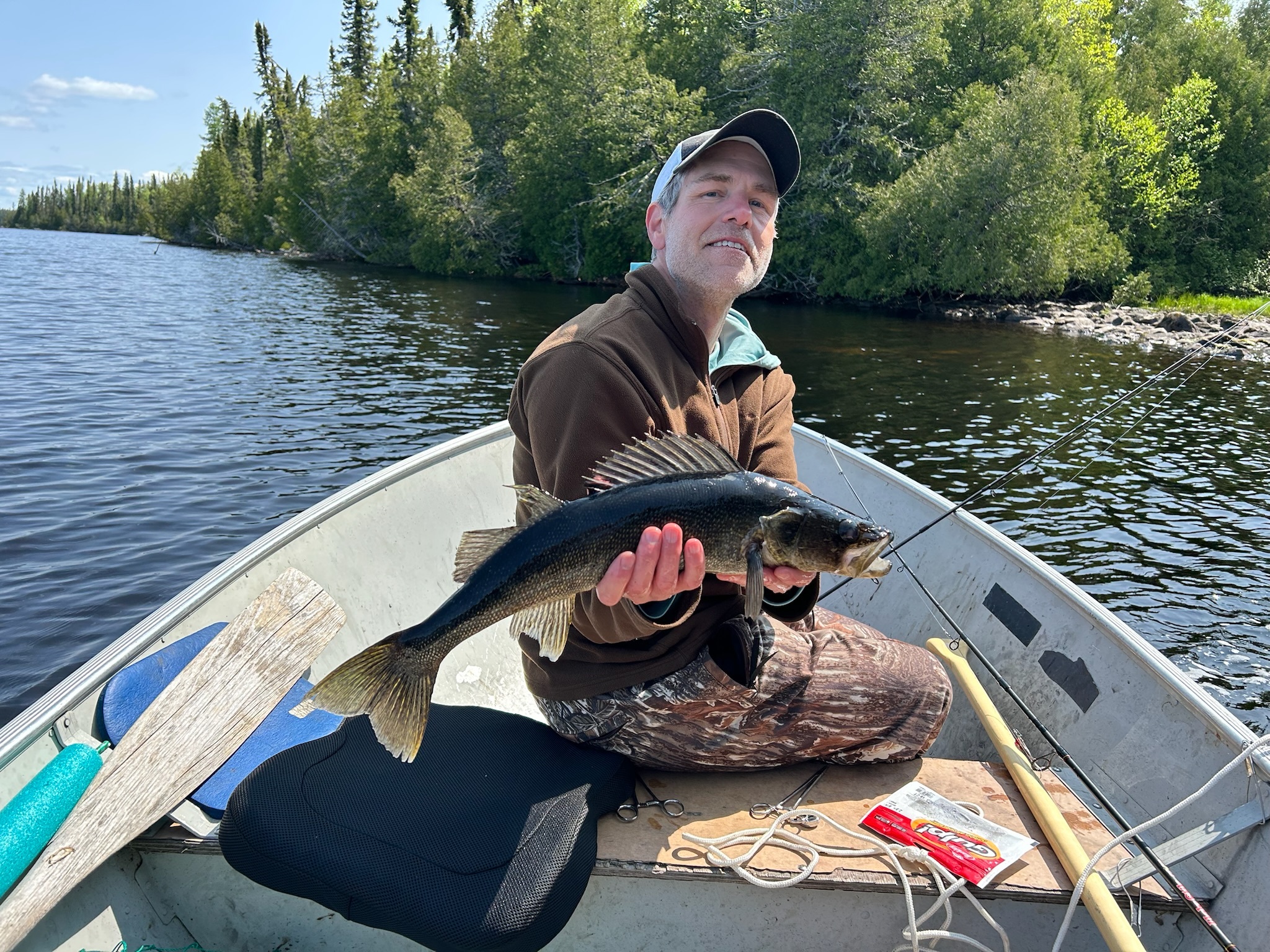 A fisherman in a small boat holds up a large walleye caught on a remote Canadian lake surrounded by dense forest.