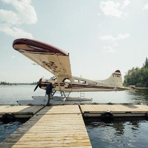 A white and maroon floatplane docked at a wooden pier on a calm lake surrounded by evergreen trees.