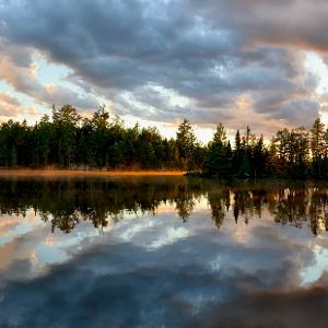 Panoramic view of a calm Canadian lake at sunset with dramatic clouds and a pine forest reflected in the still water.