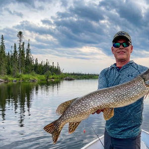 A fisherman holds a large Northern Pike horizontally in a boat on a calm lake with a forested shoreline.