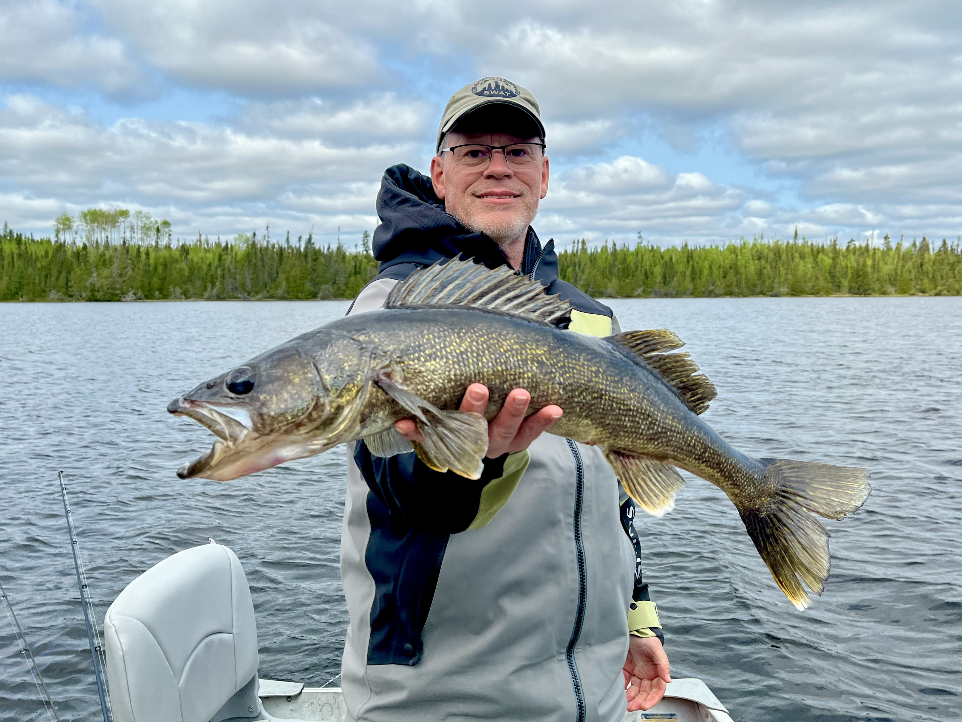 A smiling fisherman in a boat holding a large, golden walleye against a backdrop of a boreal forest and cloudy sky.